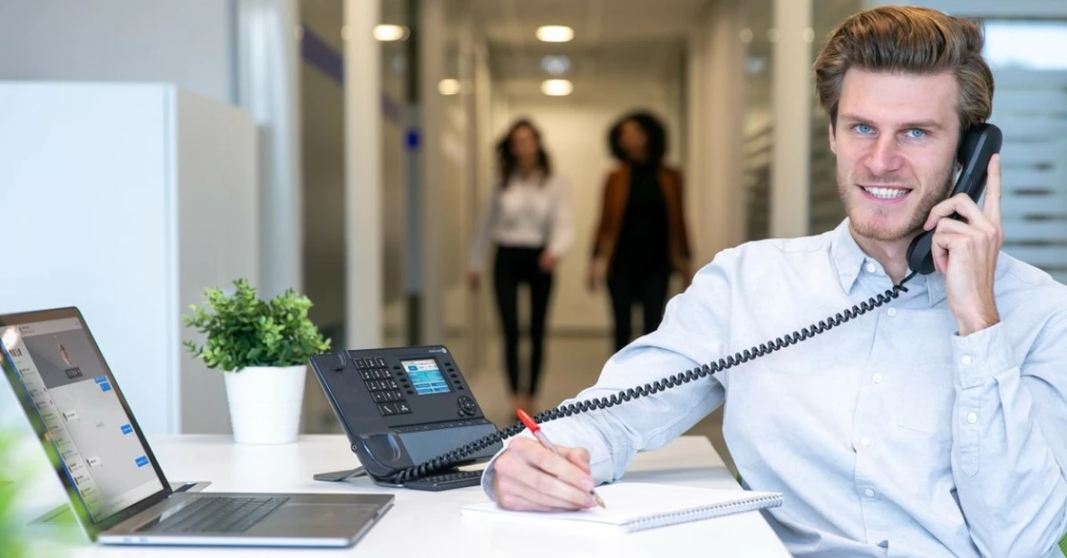 Ein Mann am Arbeitsplatz während eines Telefonats mit einem Tischtelefon von Alcatel-Lucent schaut in die Kamera