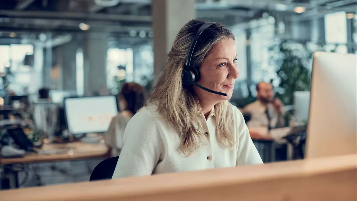 Frau sitzt mit Headset am Arbeitsplatz und lächelt